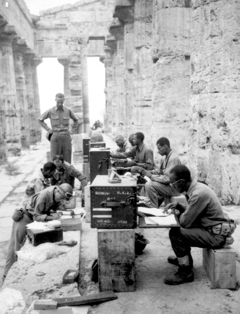 Men of the Headquarters Company, 480th Port Battalion (an African-American unit) in makeshift Fifth Army office in ancient Temple of Neptune in Paestum, Italy, 22 September 1943; At desk, front to rear: Sgts. James Shellman, Gilbert A. Terry, John W. Phoenix, Curtis A. Richardson, and Leslie B. Wood. In front of desk, front to rear: T/Sgt. Gordon A. Scott, M/Sgt. Walter C. Jackson, Sgt. David D. Jones, and WO Carlyle M. Tucker (US National Archives: 111-SC-181588)