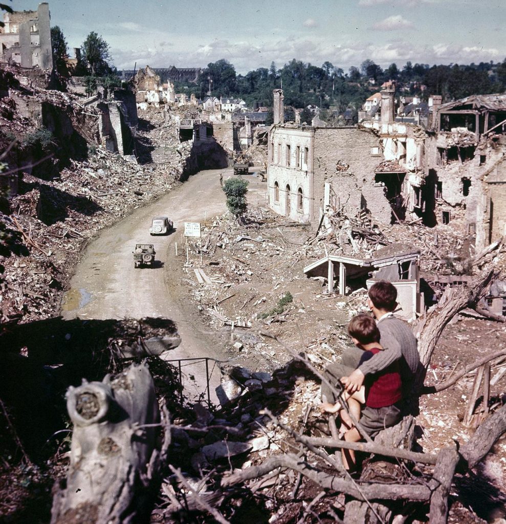 Two French boys watch as Allied vehicles pass through the ruins of Saint-Lô, France, July-August 1944 (US National Archives)